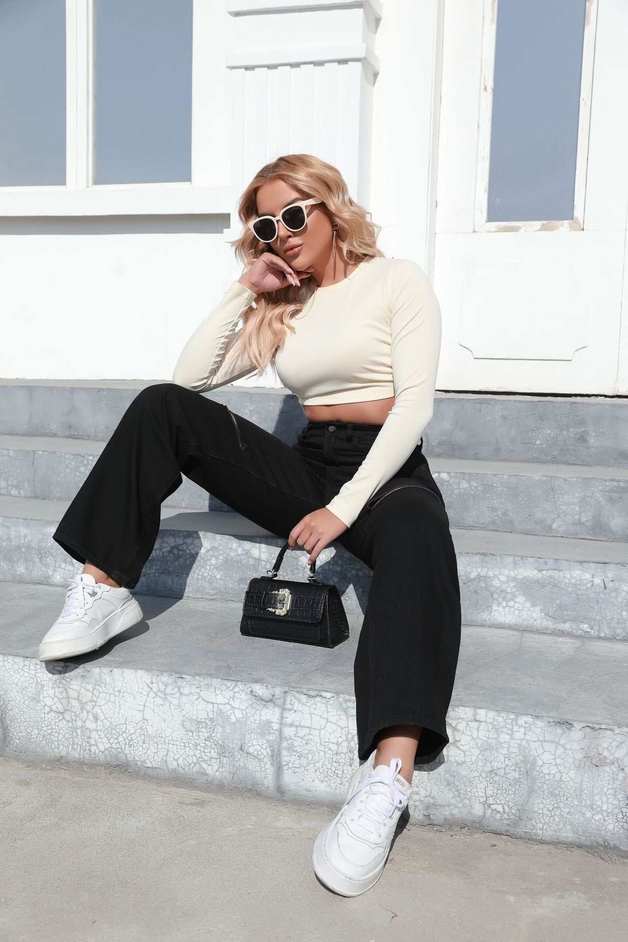 Woman wearing elegant black ripped wide-leg jeans with white sneakers and cream long-sleeve crop top sitting on outdoor concrete steps